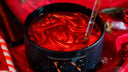 A close-up of a black candle with a glossy red wax surface and a clear glass rod inserted into it, set against a red fabric background with holiday decorations.