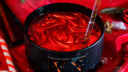 A close-up of a black candle with a glossy red wax surface and a clear glass rod inserted into it, set against a red fabric background with holiday decorations.