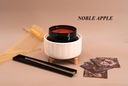 A product display featuring a white ribbed ceramic warmer with a black wickless containing a red wax melt, set on a beige background with a gold tulle bag, a black tray with a gold-toned accessory, and several cards. The brand name 'NOBLE APPLE' is printed in black text in the upper right.