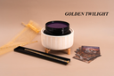 A product display featuring a white ribbed ceramic warmer with a black wickless containing a purple wax melt, set on a beige background with a gold tulle bag, a black tray with a gold-toned accessory, and several cards. The brand name 'GOLDEN TWILIGHT' is printed in black text in the upper right.
