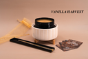 A product display featuring a white ribbed ceramic warmer with a black wickless containing a gold wax melt, set on a beige background with a gold tulle bag, a black tray with a gold-toned accessory, and several cards. The brand name 'VANILLA HARVEST' is printed in black text in the upper right.