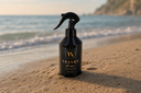 Black spray bottle labeled 'Velvet Home' on a sandy beach with ocean waves in the background