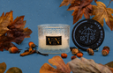 An Autumn Rain Velvet Noble candle on a blue background with decorative fall elements.