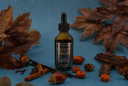 A bottle of Velvet Drops Luxury Warmer Oil centered on a textured blue background, surrounded by autumn props including brown leaves, acorns, cinnamon sticks, and a small pumpkin. The bottle is amber glass with a black dropper cap and a dark label with gold lettering.
