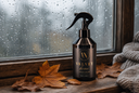 A dark velvet home spray bottle with a black trigger sprayer, set on a rustic wooden windowsill with autumn leaves and a knitted blanket nearby, with raindrops on the window in the background.