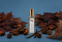 A clear glass bottle with a gold cap and a white label reading 'The Velvet Noble Autumn Rain' stands centered on a blue surface surrounded by autumn leaves, cinnamon sticks, pinecones, and vanilla beans.