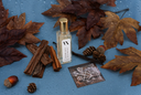 A clear glass bottle with a gold cap and a white label reading 'The Velvet Noble Autumn Rain' stands centered on a blue surface surrounded by autumn leaves, cinnamon sticks, pinecones, and vanilla beans.