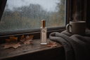 A small clear glass bottle with a gold cap stands on a rustic wooden windowsill. The bottle is labeled 'VN' and 'Autumn Rain'.