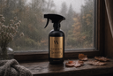 A black spray bottle with a gold label reading 'Velvet Sparkle' sits on a rustic wooden windowsill. Raindrops are visible on the window behind it, with dried autumn leaves and a knitted blanket nearby.