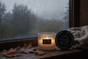 A lit candle in a textured glass jar sits on a wooden windowsill with raindrops outside, next to a round black lid and a knitted beige throw blanket.