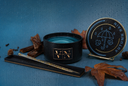 A black scented candle jar with a blue wax fill, shown beside its matching round lid on a small stand, with a stirring wand in a black holder tray. The scene includes autumn leaves, cinnamon sticks, and pinecones on a textured blue background.