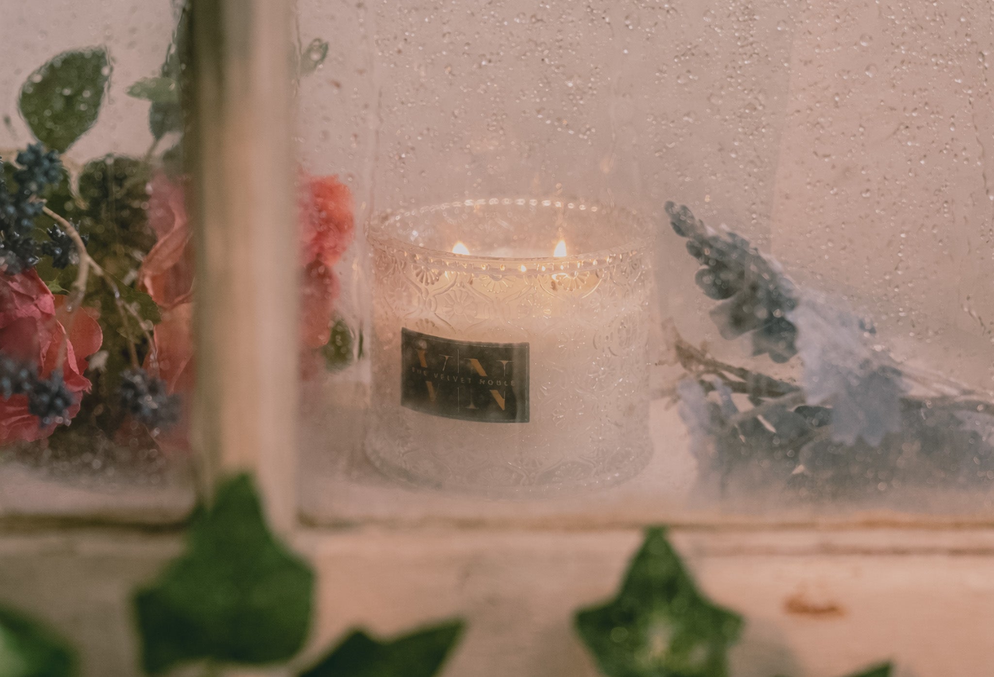 A lit candle through a wet window surrounded by flowers and greenery.