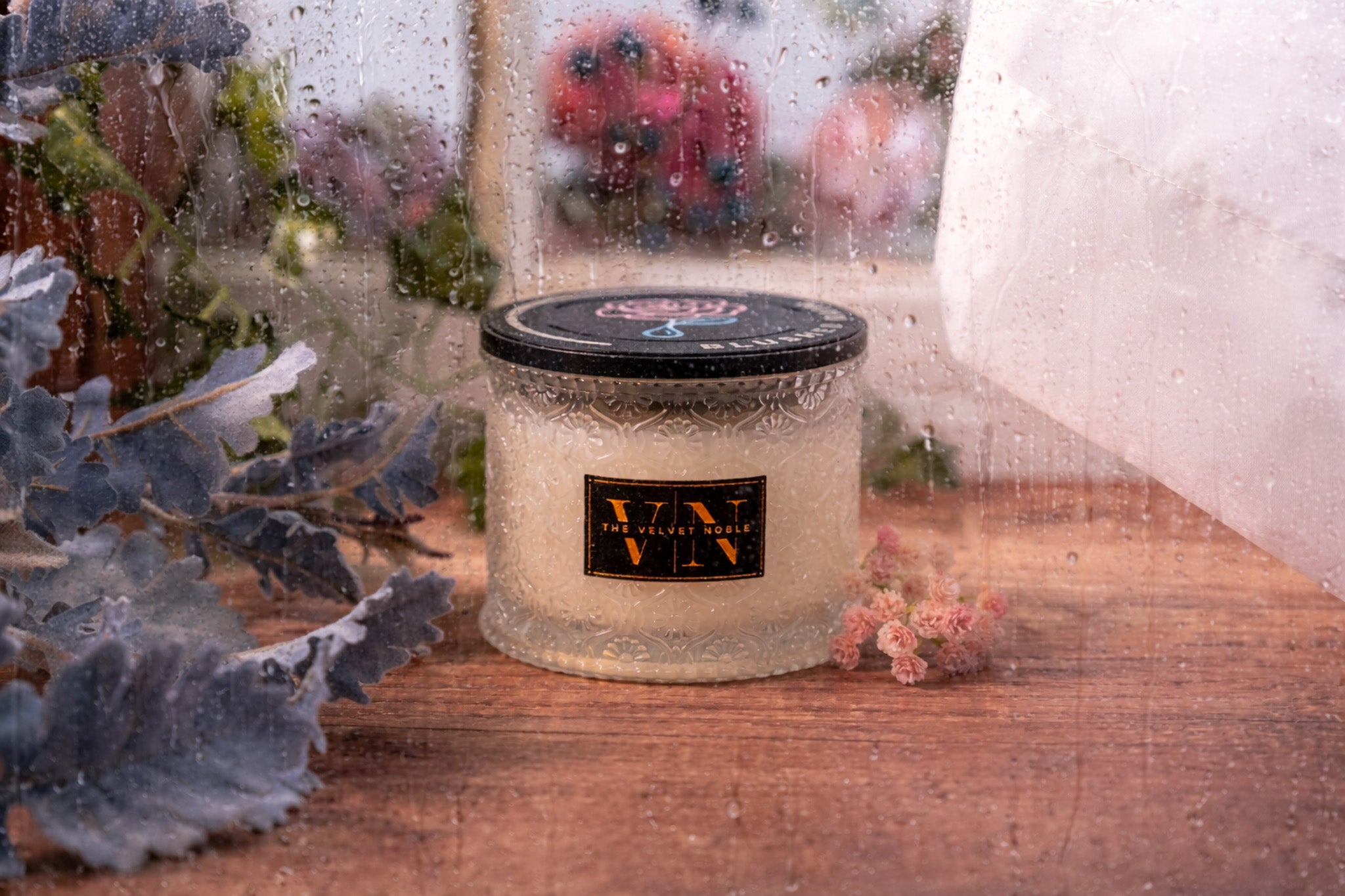 A blushed rain candle on a wet wooden surface with flowers and greenery. The frame is wet with water droplets.