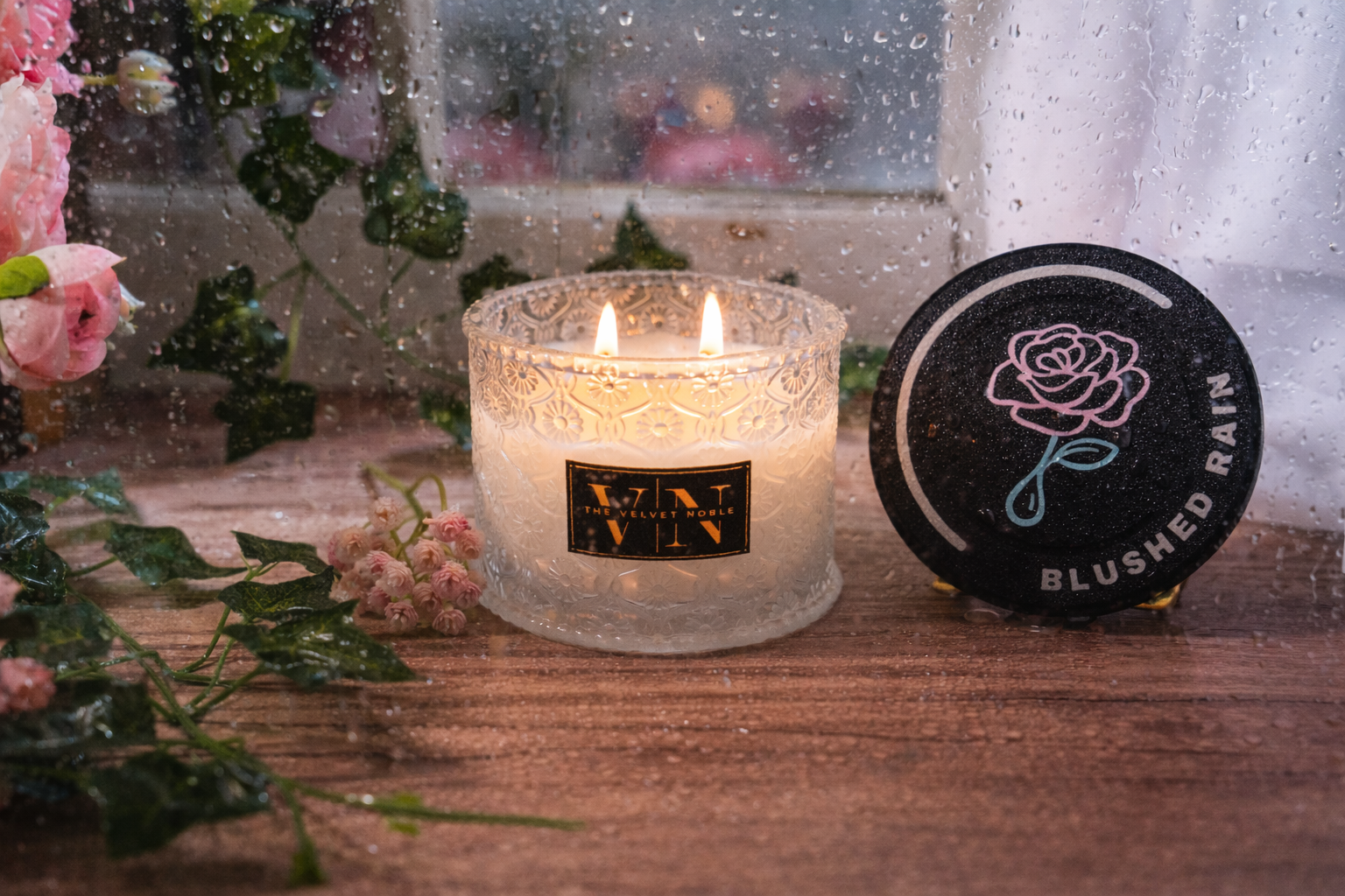A blushed rain candle lit on a wet wooden surface with flowers and greenery. The frame is wet with water droplets.