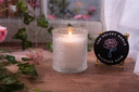 A single wick blushed rain candle on a wet wooden surface surrounded by flowers and greenery
