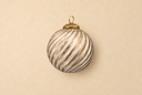 A spherical Christmas ornament with a textured, ribbed surface featuring diagonal stripes of metallic gold and distressed white, complete with a small gold hanging loop at the top, set against a solid beige background.