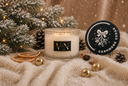 A frosted glass jar candle with a black and gold label reading 'VN' sits on a cream knit blanket beside a round black branded coaster that says 'CASHMERE CEDAR.' The scene is styled with pine branches, dried orange slices, gold bells, pinecones, and falling snow in a warm holiday setting.