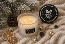 A lit double-wick candle in a frosted glass jar sits on a cream knit blanket with holiday props including pine branches, a pinecone, gold bells, dried orange slices, and a round black product lid labeled 'CASHMERE CEDAR' and 'VN'.