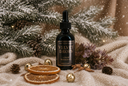 A dark amber glass dropper bottle labeled 'VELVET DROPS' by 'THE VELVET NOBLE' sits on a cream knit blanket with pine branches, dried orange slices, gold bells, and pinecones in a festive holiday setting.