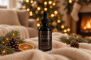 A bottle of The Sacred Purpose Velvet Drops Luxury Warmer Oil stands on a cream knit blanket with Christmas pine branches, pinecones, and a dried orange slice, with a blurred Christmas tree and fireplace in the background.