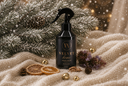 A black spray bottle labeled 'VELVET home' with gold lettering, styled on a beige knit blanket with pine branches, dried orange slices, gold bells, and soft holiday lights in the background.