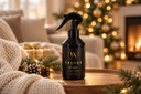 A black trigger spray bottle labeled 'VELVET home' stands on a wooden table in a cozy holiday living room setting with a fireplace, warm lights, pine branches, and a knitted blanket.