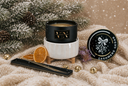 A black scented candle on a white ribbed holder warmer on a beige knit blanket with winter-themed props, including pine branches, dried orange slices, small gold bells, and a round black lid labeled 'CASHMERE CEDAR'.