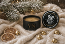 A black candle tin and its lid are arranged on a cream knit blanket with holiday decor, including pine branches with snow, dried orange slices, small gold bells, and warm string lights. The lid reads 'CASHMERE CEDAR' and the tin shows 'VN' branding.