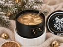 A black jar candle with a gold label reading 'VIN The Velvet Room' sits open on a white pedestal, filled with a swirled gold wax. A gold glittery wick extends upward.