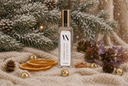 A clear rectangular bottle of Cashmere Cedar Air & Linen Mist with a gold cap and white label, styled on a beige knit blanket with dried orange slices, gold bells, pinecones, and winter greenery in the background.