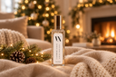 A clear glass bottle of Cashmere Cedar Air & Linen Mist with a gold cap and white label, set on a cozy beige knit blanket with pine branches and a warm fireplace background.