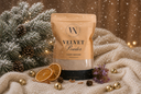 A stand-up pouch of Velvet Powder luxury freshener stands on a cream knit blanket with holiday props including frosted pine branches, dried orange slices, small gold bells, and a sprig of purple flowers. The pouch is tan with black text and a black base, photographed in a warm festive setting with soft lights and falling snow effects.
