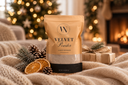 A stand-up pouch of Velvet Powder luxury freshener by The Velvet Company, styled on a cozy beige knit blanket with pinecones, dried orange slices, and wrapped gifts, with a blurred Christmas tree and fireplace in the background.