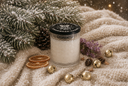 A festive white candle in a textured glass jar with a black lid, styled on a cream knit blanket with pine branches, gold bells, dried orange slices, and purple flowers.