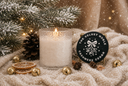A lit white candle in a textured glass holder sits on a cream knit blanket beside a round black product tag reading 'THE VELVET NOBLE CASHMERE CEDAR.' The scene is styled with pine branches, dried orange slices, small gold bells, pinecones, and falling snowflake effects, creating a cozy holiday atmosphere.