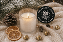 A lit white candle in a textured glass holder sits on a snowy surface beside a round black product label reading 'THE VELVET NOBLE CASHMERE CEDAR.' The scene is styled with pine branches, pinecones, dried orange slices, small gold bells, and soft snow, creating a cozy holiday atmosphere.