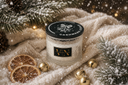 A glass jar candle labeled 'CASHMERE CEDAR' by 'The Velvet Noble' sits on a cream knit blanket surrounded by pine branches, dried orange slices, gold bells, and falling snow-like confetti.