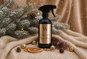A black trigger spray bottle with a gold label reading 'Velvet Sparkle' stands on a beige knit blanket surrounded by holiday props including pine branches, gold bells, dried orange slices, and a small sprig, against a warm draped fabric backdrop with falling snowflake overlay.