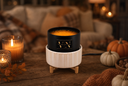 A scented wickless candle in a ribbed white ceramic warmer with small wooden legs, set on a rustic wooden table in a cozy autumn living room. The candle is surrounded by lit candles, pumpkins, fall leaves, and a knitted blanket.