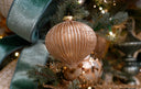 A champagne colored teardrop shaped, ribbed glittered ornament, on a green Christmas free, with other gold and green accents.