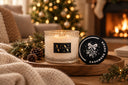 A lit scented candle in a textured glass jar sits on a wooden tray beside its black lid in a cozy living room setting with a fireplace, Christmas tree lights, and a knitted blanket.
