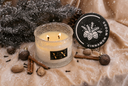 A lit candle in a frosted glass jar with two wicks sits on a beige fabric surface with cinnamon sticks, nutmegs, pinecones, and a black round decorative lid printed with the scent name CINNAMON WOODS.