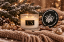 A festive holiday candle set styled on a knitted beige blanket with snow-dusted pine branches, warm string lights, cinnamon sticks, and gold ornaments. The set includes a frosted glass candle jar with a black and gold label reading 'VN' and a round black lid with white pinecone artwork and the words 'CINNAMON WOODS'.