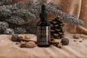 A dark amber glass dropper bottle labeled 'The Velvet Noble Velvet Drops Luxury Warmer Oil' styled on a beige fabric surface with cinnamon sticks, walnuts, pinecones, and evergreen branches, with a snowy, festive background.