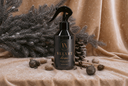A black spray bottle labeled 'VELVET home' stands on a beige fabric surface with decorative pine branches, pinecones, and small ornaments. The background is a draped beige fabric with a soft, warm tone.