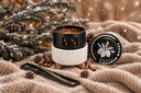 A wickless candle product displayed on a cozy beige knit blanket with snowy pine branches, pinecones, cinnamon sticks, and warm string lights in the background. The candle jar is black with a cream ribbed warmer and a gold label reading 'The Velvet Noble', next to its matching lid labeled 'CINNAMON WOODS'.