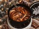 A close-up of a black candle jar filled with a glossy, swirled copper-brown wax, with a glass stirring wand resting in the wax. The scene is styled with a snowy holiday theme, including pine branches, cinnamon sticks, and a snow-dusted lid in the background.