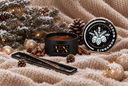 A black candle jar labeled 'VN Cinnamon Woods' and its matching lid are styled on a beige knit blanket with pine branches, cinnamon sticks, pinecones, and gold ornaments. The product appears to be a candle, with the lid shown alongside the open jar.