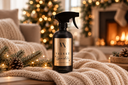 A black trigger spray bottle labeled 'Velvet Sparkle' stands on a cream knit blanket in a cozy holiday living room with a fireplace, Christmas tree lights, and pine branches.