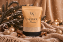 A stand-up pouch of Velvet Powder luxury freshener by The Velvet Noble, styled on a cozy beige knit blanket with cinnamon sticks, pinecones, and gold holiday ornaments against a warm draped backdrop.